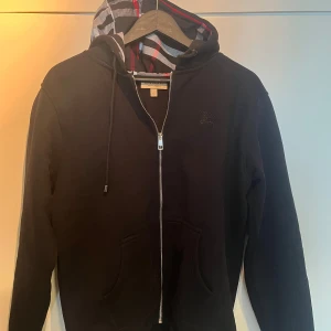 Svart hoodie från Burberry - Säljer en snygg svart hoodie från Burberry. Den har en dragkedja framtill och en klassisk huva med ett mönstrat foder. Perfekt för höst och vinter, den är både stilren och bekväm. Passar till alla tillfällen. 9/10 skick.  BVSA!