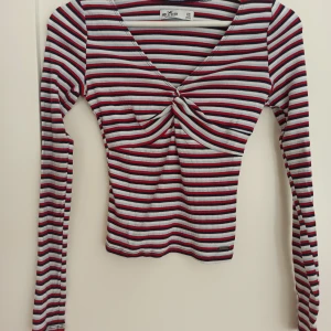 Randig långärmad topp från Hollister - Säljer en superfin randig topp från Hollister i storlek XS. Den är långärmad och har en snygg knyt-detalj framtill. Färgerna är röd, vit och blå, och materialet är ribbad bomull. Perfekt för både vardag och fest! 💖 Aldrig använd av mig, skick som ny! Verkligen såå fin passform🌟