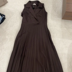 Brun ärmlös klänning - Säljer en elegant brun ärmlös klänning med veckad kjol. Klänningen har en klassisk krage och omlott-design upptill, vilket ger en stilren look. Perfekt för både fest och mer formella tillfällen.