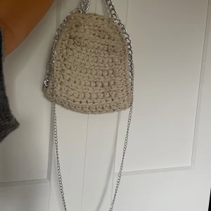 Beige stickad axelväska med kedjedetaljer - Säljer en superfin beige stickad axelväska med coola kedjedetaljer. Perfekt för att ge din outfit en unik touch! Väskan är handgjord och har en snygg, bohemisk stil. Den är både praktisk och trendig, perfekt för vardagsbruk eller en dag på stan.