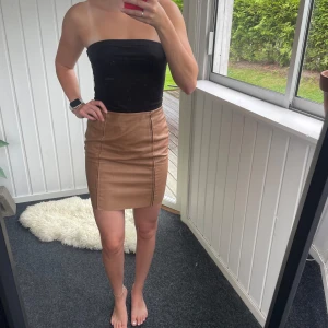 Brun mockakjol från Pieces - Brun ”skinnkjol” från Pieces i storlek S. Kjolen är tight, kort och i syntetmaterial. Dragkedja i sidan. Defekter förekommer (se bild).