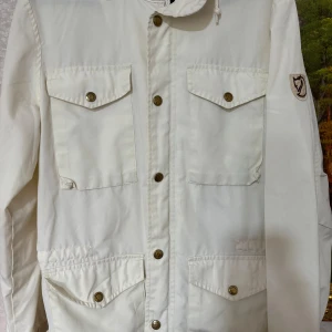 Vit jacka från Fjällräven - Säljer en snygg vit jacka från Fjällräven. Den har en klassisk design med fyra fickor framtill och knappar. Perfekt för vår och höst!