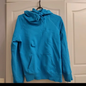 Blå hoodie - Säljer en snygg blå hoodie i bra skick. Den har en stor ficka fram och en justerbar huva med snören. Perfekt för kyliga dagar eller mysiga hemmakvällar. Passar både till vardags och avslappnade tillfällen.