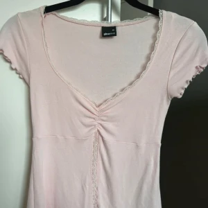 Ljusrosa topp från Gina Tricot - Säljer en supersöt ljusrosa topp från Gina Tricot i storlek XS. Toppen har korta ärmar och en v-ringad design med spetsdetaljer längs kanterna. Den har en fin rynkning framtill som ger en extra touch. Knappt använd.