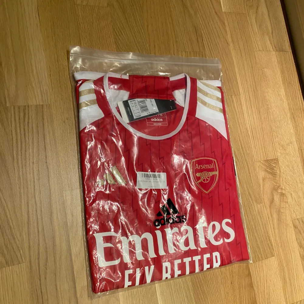 Säljer en ny Arsenal fotbollströja i rött och vitt från Adidas. Tröjan har korta ärmar och ett snyggt mönster med klubbens emblem och sponsorlogga på framsidan. Storlek M & perfekt för alla Arsenal-fans! 🏆. T-shirts.