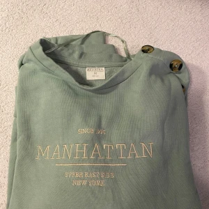 Grön sweatshirt från & Other Stories - Säljer en snygg grön sweatshirt från & Other Stories med texten 'Manhattan Upper East Side New York' på bröstet. Tröjan har långa ärmar och coola knappar på axeln. Perfekt för en avslappnad stil! 💚