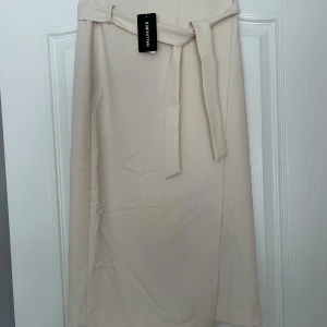 Krämfärgad kjol från Hallhuber - Säljer en elegant krämfärgad kjol från Hallhuber i storlek 36. Kjolen har en stilren design med ett knytband i midjan för en justerbar passform. Perfekt för både vardag och festliga tillfällen. Materialet känns mjukt och bekvämt mot huden.