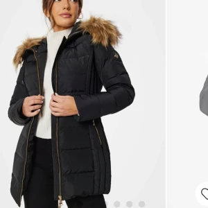 Svart dunjacka med päls - Snygg svart dunjacka från Hollies med päls på huvan. Jackan har en dragkedja framtill och är perfekt för kalla vinterdagar. Den är långärmad och quiltad för extra värme. Perfekt för både vardag och festliga tillfällen!