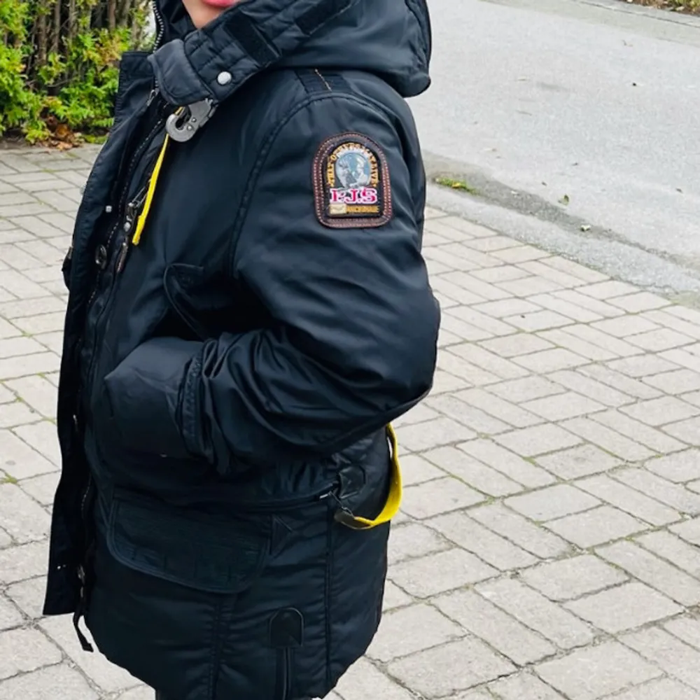 Säljer en snygg och varm mörkblå parkajacka från Parajumpers. Jackan har en praktisk huva och långa ärmar, perfekt för kalla vinterdagar. Den är utrustad med dragkedja och knappar för extra skydd mot kylan. Märkesloggan syns på ärmen, vilket ger en cool touch. Passar perfekt för både vardag och utomhusaktiviteter.. Jackor.