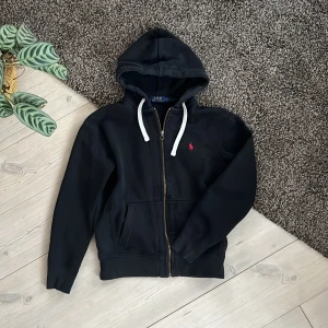 Ralph Lauren Zip Hoodie - Tjena, jag säljer denna snygga zip hoodie från Ralph Lauren i storlek S. Den är i 100% bomull och man kan verkligen känna den fina kvaliteten på den tunga vikten och det tjocka Hoodien är i bra skick. Jag har sytt upp ett litet hål vid fickan tidigare men det är inget som man märker och ingen annan skulle märka det heller. Ställ gärna frågor!