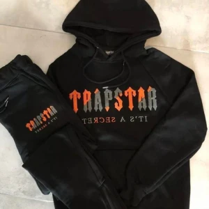 Svart hoodie från Trapstar - Snygg svart dress från Trapstar med tryck i orange och grått på bröstet. Perfekt för en casual look med en skön passform och klassisk huva. Intresserad! Skriv till mig på DM