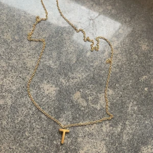 Guldhalsband med bokstav - Elegant och personligt guldhalsband med bokstaven T. Kedjan är tunn och smidig med en liten märkestagg vid låset. Perfekt för en stilren look. Knappt använd🫶🏼