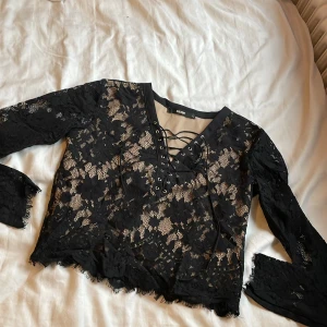 Svart spetsblus med snörning - Snygg svart spetsblus från bikbok med långärmad design och snörning framtill. Perfekt för en trendig look med sina fina spetsdetaljer och v-ringad hals. Blusen är croppad och har en elegant stil. Använd två gånger