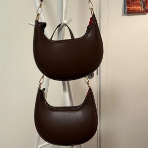 Brun axelväska i skinn - Snygg brun axelväska i skinn med en stilren design. Väskan har en rundad form och justerbar axelrem i samma material. Perfekt för att bära dina dagliga nödvändigheter med stil. 50kr för 1 eller båda för 100!