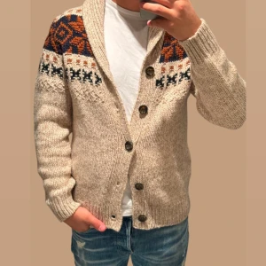 Beige stickad kofta med mönster - Säljer en mysig beige stickad kofta med ett färgglatt mönster i blått och orange över axlarna. Koftan har knappar framtill och långa ärmar, perfekt för kyliga dagar.