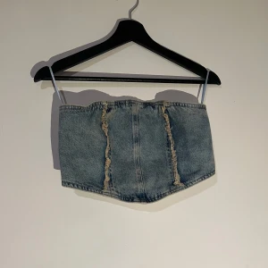 Jeans korsett bandeau top - Trendig jeans korsett bandeau med knappar framtill och råa kanter. Perfekt för att ge en edgy touch till din outfit. Den har en klassisk blå denimfärg och är designad för att sitta tight.
