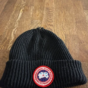Svart mössa Canada Go*se - Snygg svart ribbad mössa från Canada Goose med det ikoniska Arctic Program-emblemet framtill. Perfekt för kalla dagar och ger en stilren look.