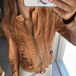 Äkta Brun skinnjacka från Rock’n Blue - Snygg brun skinnjacka med dragkedja och ståkrage. Jackan har långa ärmar och tryckknappar vid kragen och ärmsluten. Perfekt för en stilren look. Liten i storleken, nypris ca 2700kr