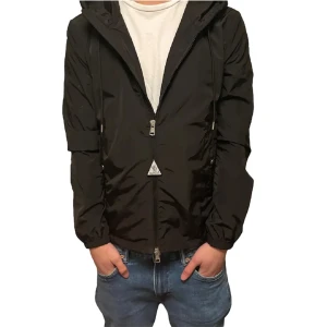 Moncler windbreaker  - Snygg svart vindjacka från Moncler med dragkedja och huva. Jackan har ett broderat Moncler-märke på ärmen och ränder i rött och vitt på huvan. Perfekt för blåsiga dagar. Inget og medföljer