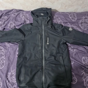 Svart jacka från Tenson - Säljer en svart jacka från Tenson med dragkedja och huva. Jackan är vind- och vattentät med andningsfunktion, perfekt för utomhusaktiviteter. Den har en stilren design med Tenson-logga på bröstet och armen