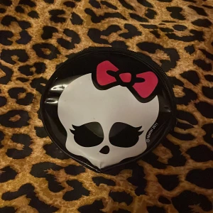 Monster Hugh plånbok - Plånbok/miniväska med ett dödskallemotiv i vitt och en rosa rosett. Perfekt för att förvara småsaker. Den har en praktisk dragkedja och är lätt att bära med sig.
