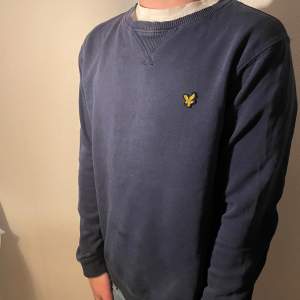 Säljer en stilren mörkblå tröja från Lyle & Scott med deras ikoniska logga på bröstet. Tröjan har långa ärmar och en rund halsringning med ribbade detaljer. Perfekt för en avslappnad look.
