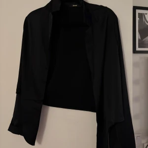 Svart blus från bikbok - Elegant svart blus från bikbok  med en klassisk design. Perfekt för en stilren look. Blusen har långa ärmar och en öppen front utan knappar.