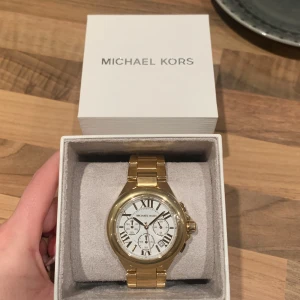 Guldfärgad klocka från Michael Kors - Helt ny guldfärgad klocka från Michael Kors med vit urtavla och romerska siffror. Klockan har tre mindre urtavlor och en robust länkarmband i metall. Perfekt för att ge en lyxig touch till din stil. Plasten är kvar, nypris ca 3500kr