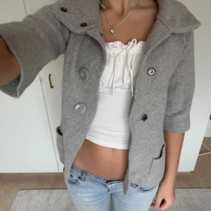 Grå stickad kappa med stora knappar - Säljer en stilren grå stickad kappa med stora knappar och trekvartsärm. Perfekt för höst och vår, med en mysig krage och praktiska fickor framtill. Enkelt att matcha med olika outfits!