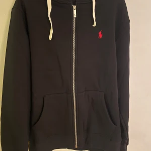 Svart hoodie från Ralph Lauren - Säljer en helt oanvänd svart hoodie från Ralph Lauren med dragkedja och röd logga på bröstet. Storlek L men passar M