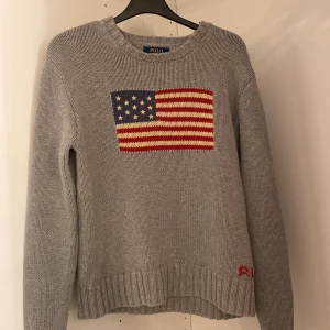 Grå stickad tröja från Polo Ralph Lauren - Snygg grå stickad tröja från Polo Ralph Lauren med amerikansk flagga på bröstet. Tröjan har långa ärmar och ribbstickade muddar vid ärmslut och nederkant. Perfekt för en avslappnad stil.