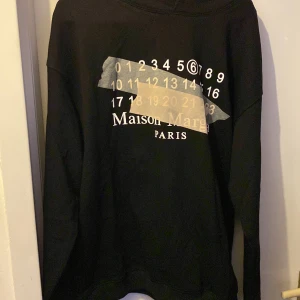 Svart hoodie från Maison Margiela - Säljer en stilren svart hoodie från Maison Margiela med deras ikoniska siffror och logga på framsidan. Tröjan har långa ärmar och en klassisk passform. Perfekt för en avslappnad och trendig look.