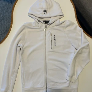 Vit hoodie från Sail Racing - Säljer en snygg vit sail racing. Det är en zip hoddie med logga på bröstet. Finns defekter lite sliten på är ärmslut och lite nopprig på insidan men inget som syns.