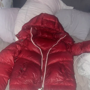 Röd dunjacka från Moncler - Säljer en snygg röd dunjacka från Moncler med dragkedja och huva. Jackan har en quiltad design och praktiska fickor framtill. Perfekt för kyliga dagar!