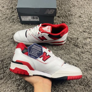 DSWT! New Balance 550 i vitt, rött och svart  - Snygga New Balance 550 sneakers i vitt med röda och svarta detaljer. Skorna har en klassisk design med snörning och en bekväm passform. Perfekta för en sportig look.