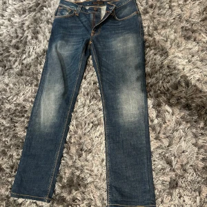 Blå nudie jeans - Säljer ett par klassiska blå jeansbyxor med en straight passform. Byxorna har en midwaist och är tillverkade i denim med snygga slitningar på framsidan. De har fem fickor och stängs med dragkedja och knapp.