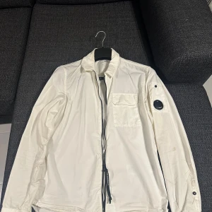 Vit jacka/ cardigan C.P. Company - Säljer en stilren vit jacka från C.P. Company med dragkedja och en unik detalj på ärmen. Jackan har en bröstficka och långa ärmar, perfekt för en modern look. Knappt använt den, har legat mycket i gaderoben. 