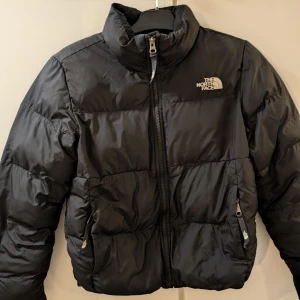 Svart pufferjacka från The North Face - Säljer en svart pufferjacka från The North Face. Jackan har en dragkedja framtill och två fickor med dragkedjor. Perfekt för kyliga dagar med sin vadderade design.