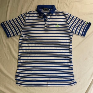 Randig pikétröja från Polo Golf Ralph Lauren - Snygg blå och vit randig pikétröja från Polo Golf Ralph Lauren med broderad logga för US Open Chambers Bay 2015. Perfekt för golfentusiasten. Tröjan har korta ärmar och en klassisk krage med knappar.