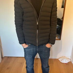 Svart/ mörkblå stone Island jacka  - Snygg svart pufferjacka från Stone Island med huva och dragkedja. Jackan har en broderad logga på ärmen och är perfekt för kyligare dagar.