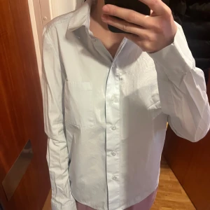 Blue skjorta från COS - Säljer en stilren vit skjorta från COS i storlek 36. Skjortan har en klassisk krage och knappar framtill. Perfekt för en minimalistisk look.