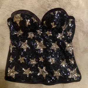 Intimissimi korsett med stjärnpaljetter - Snygg svart korsett dekorerad med glittriga stjärnformade paljetter i guld. Perfekt för en festlig kväll. Toppen har en åtsittande passform och är axelbandslös. 💗