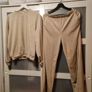 Beige sweatshirt och byxor set - Säljer ett bekvämt och mjukt set i velour. Tröjan har långa ärmar och en rund halsringning, medan byxorna har en elastisk midja för extra komfort. Perfekt för en avslappnad dag hemma. Köpt på Cubus. 