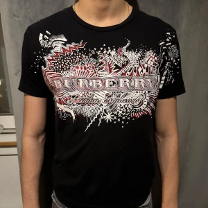 Svart t-shirt från Burberry - Snygg svart t-shirt från Burberry med ett coolt grafiskt tryck i vitt och rött. Köpt från HeatNlux för 1 år sen. Hör av er vid frågor! Tshirten har lite små pinnhål längst ner men inget som syns pga färgen