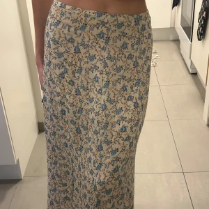 Maxi kjol - Säljer en vacker långkjol med blommigt mönster i blått och beige. Kjolen har en elegant och flytande passform, perfekt för en somrig look. Materialet känns lätt och bekvämt mot huden.