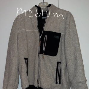 Beige fleecejacka med svarta detaljer - Säljer en mysig beige fleecejacka i storlek medium. Den har svarta detaljer på fickorna och bröstet, samt en dragkedja framtill. Perfekt för kyliga höst- och vårdagar. Jackan är i bra skick och redo för nya äventyr!