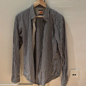 Randig skjorta från Boss Orange - Säljer en stilren randig skjorta från Boss Orange. Skjortan är långärmad och har en klassisk krage med knappar framtill. Den är i en snygg kombination av blått och vitt, perfekt för både vardag och fest. Materialet känns bekvämt och passar bra till både jeans och chinos.