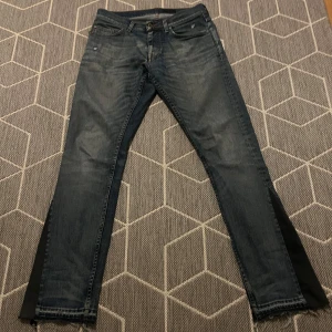 Blå jeans med svarta detaljer - Säljer ett par custommade jeans från märket ”Tiger of Sweden” med svarta paneler på sidorna som jag har gjort själv. De har en sliten look med fransiga kanter vid bensluten. Har även gjort egna slitningar. Perfekta för en avslappnad stil. Passar bra till både vardag och fest!