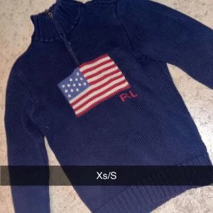 Mörkblå stickad tröja med amerikansk flagga - Säljer en snygg mörkblå stickad tröja från Ralph Lauren med en stor amerikansk flagga på bröstet. Tröjan har en dragkedja vid halsen och är perfekt för kyliga dagar. Passar både till vardags och lite finare tillfällen. Supermysig och stilren! 🇺🇸