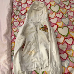 Vit ed hardy kofta  med tryck - Säljer en vit ed hardy med coolt tryck av ett hjärta och texten 'Love Kills Slowly'. Den har dragkedja och huva, perfekt för en avslappnad stil. Några små fläckar finns, men annars i bra skick eftersom den knappast syns som ni ser på tredje bilden. Står inte storleken men gissar på S-M då den sitter tajt